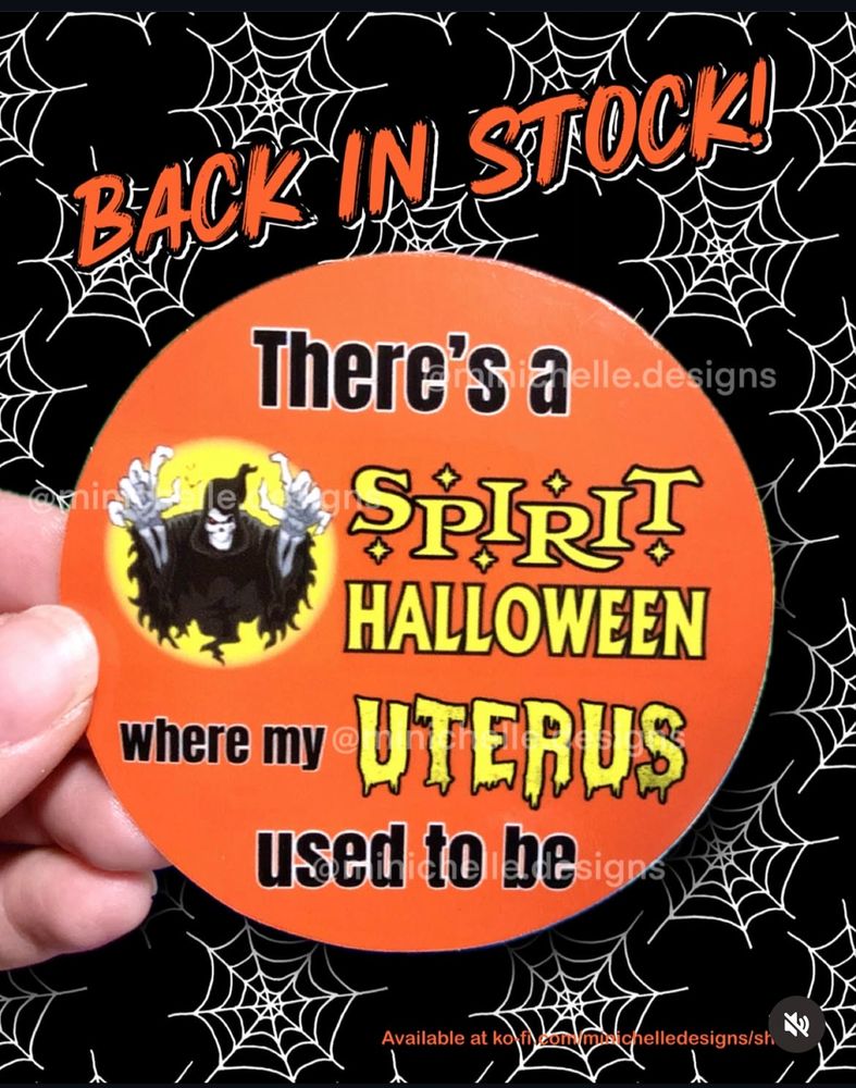 An orange circle sticker that says There’s a spirit Halloween where my uterus used to be on top of a black background with white spiderwebs