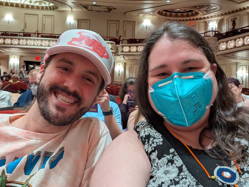 Left is a man with a brown beard wearing a hat and pastel pink shirt. Right is a brunette with a teal N95 and a black and white flowered top with a cool necklace from Kenya!!!