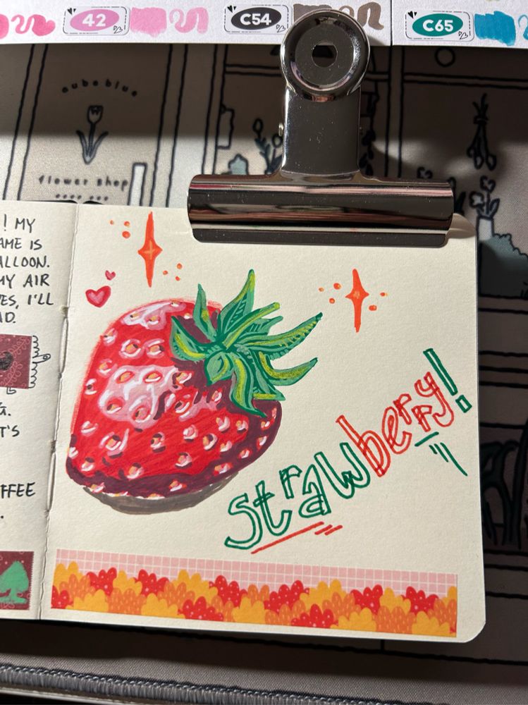 A red strawberry drawn with acrylic markers. It has green leaves on top. The page also has the word strawberry on it written with normal marker. The word straw is green and berry is red. There’s also washitape on the page, with an illustration of bushes coloured red, orange and yellow presenting autumn.