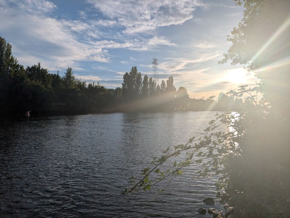 A photograph of the same channel of water, lined in the distance by trees and a blue sky, this time catching the sun as it sinks toward the water, a few hours from dusk. The sun is streaky, a product of a dirty camera lens.