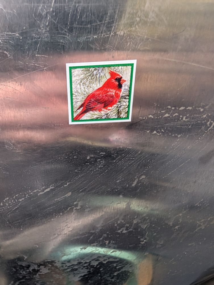 A tiny square sticker on the back of a dirty but reflective bus seat. The sticker depicts the side view of a male cardinal perched on snowy evergreen branches.
