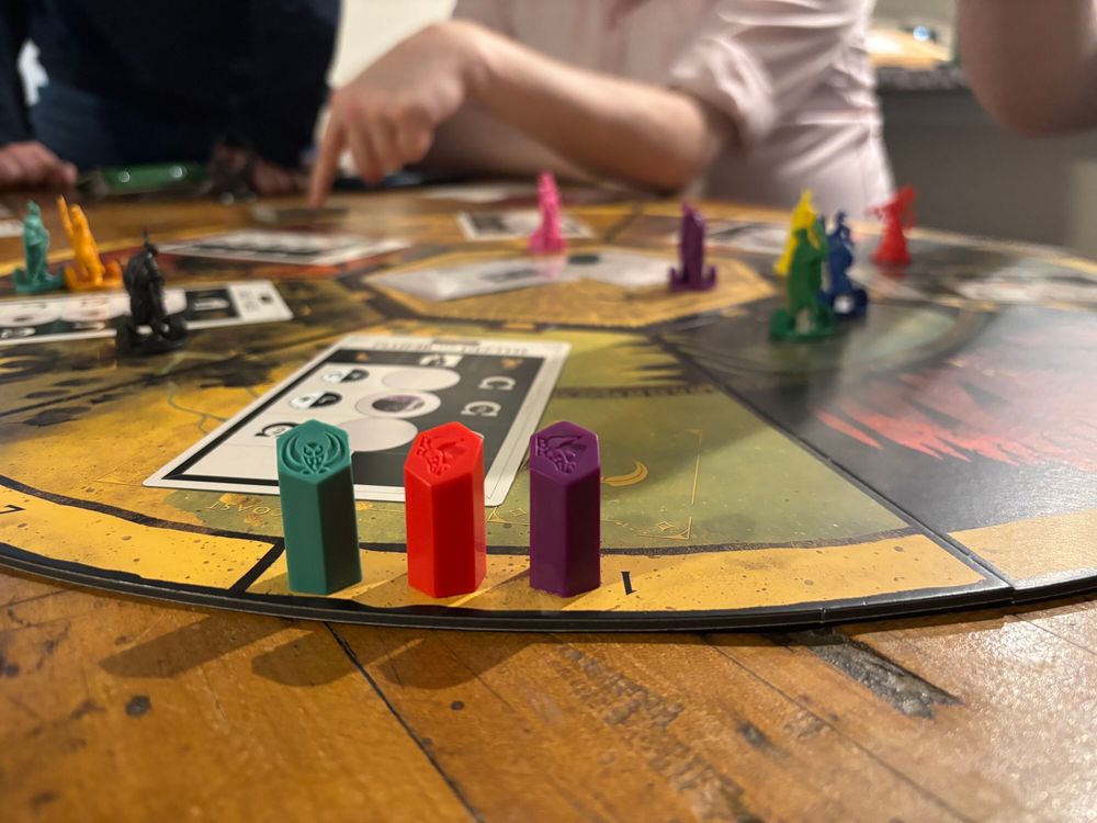 Seven players mid-game of Veiled Fate, leaning in with intense focus. A Blue player quietly smiles as scores are tallied—victory through secrecy.