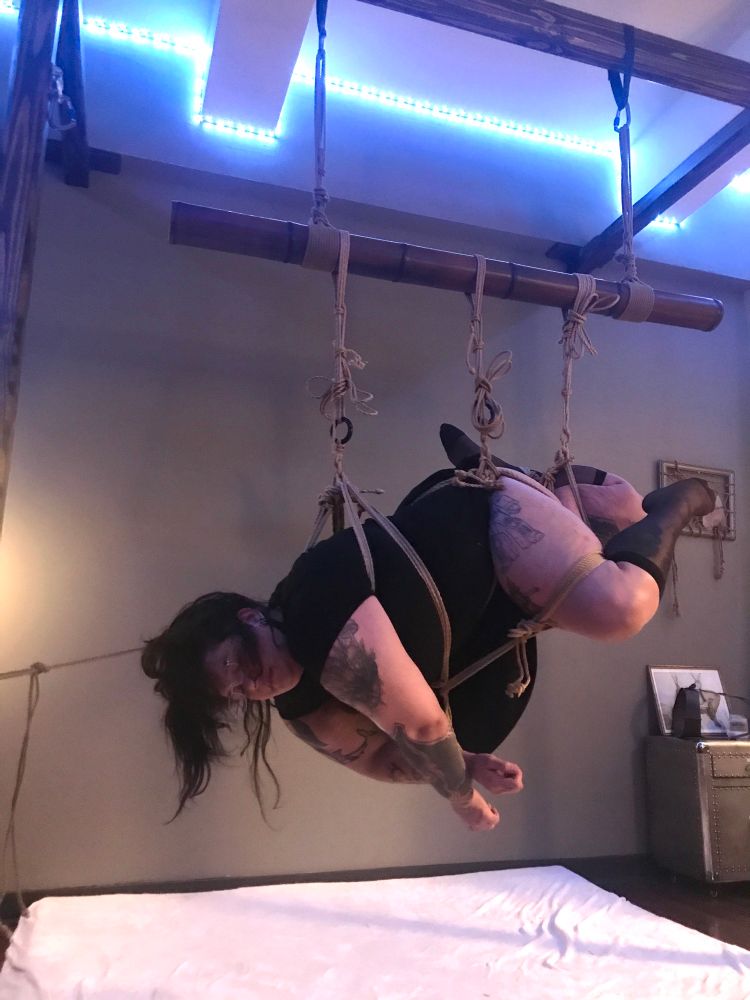 The same suspension. The line from her thigh cuff used to hold her hands together, and they are tied into the front of her chest harness. Her hair rope is tied to the side of the suspension rig.