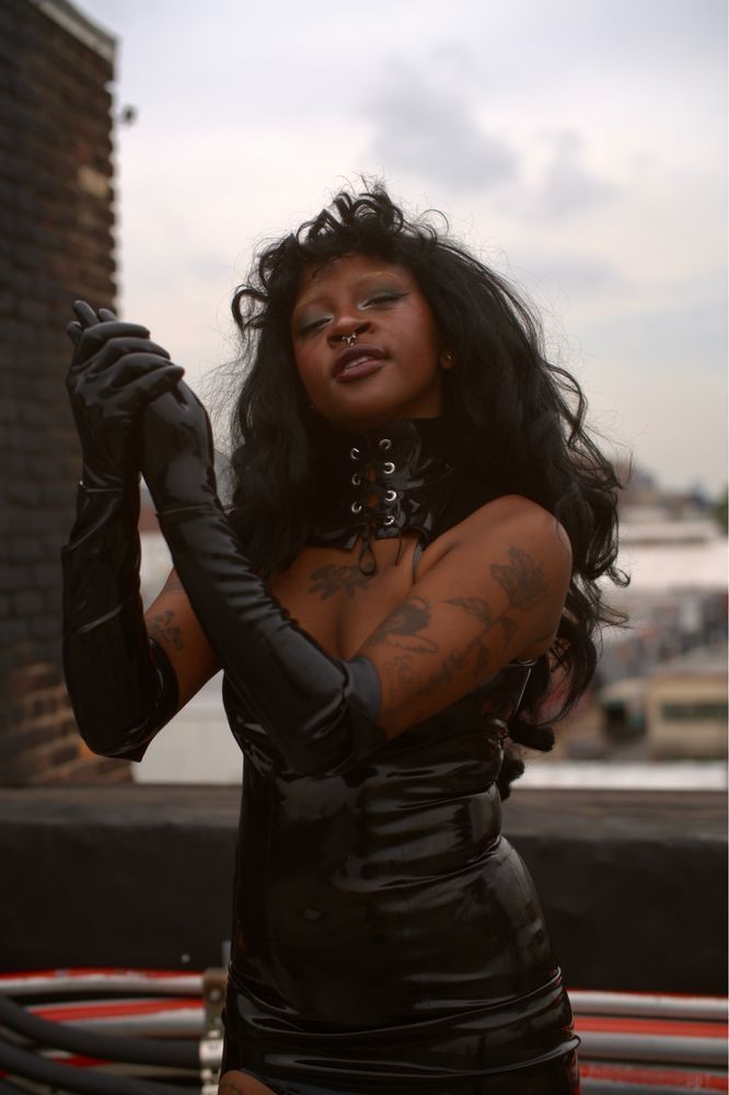 Mistrix Salem applies silicone lubricant to their hand, which is in a latex glove. they are wearing an all black latex outfit, looking into the camera with deep lust in their eyes.