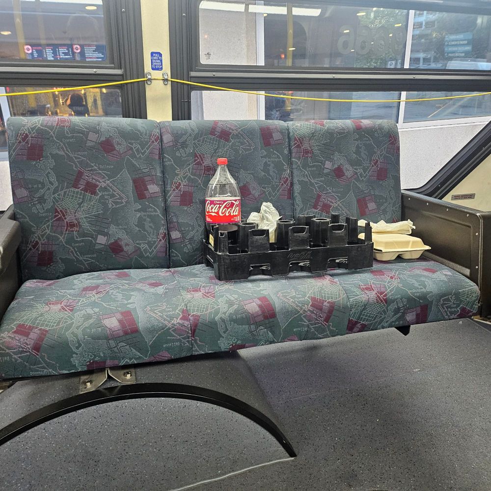 A half full 2 literally cherry cola in a plastic device built to hold 8 2 liter bottles. Also has a plastic bag and a takeout container with it, all on a wide bus seat.