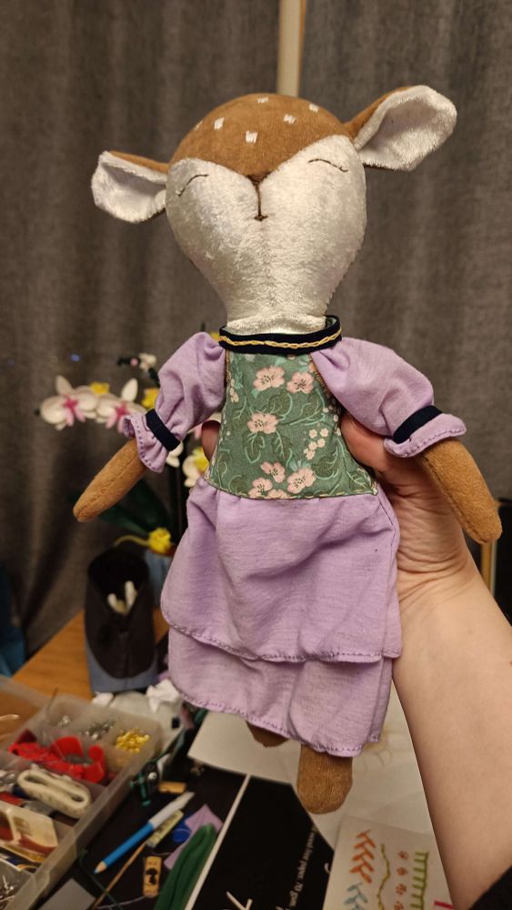 A photograph of a stuffed animal. It's a fawn. The design is basically doll style, so it's not standing on all fours, but rather has a slender, humanoid body shape, though the head is comparatively big. The body is made up of a soft white and light brown velvet, sourced ages ago from a bag of random cut-offs I bought from a theatre via Ebay. Six white dots have been embroidered on the fawn's forehad, and a closed pair of eyes, a nose and a small mouth have been embroidered on the lower, white part of the face.

The dress has a green bodice with pink and white flowers, while the shirt and sleeves are both made from the same wavey, purple fabric. The sleeves are tightened around the arm about halfway down with a dark blue band, and there's a bit of purple fabric left to flare out beyond that. The same dark blue is used for the inside of the bodice (not visible) and the horizontal neck part.