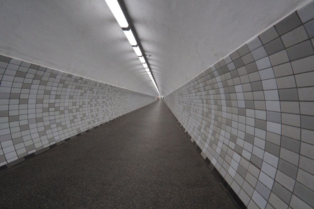 Runder, langer Tunnel, dunkler Boden, Leuchtstoffröhren an der weißen Decke,die Seiten in verschiedenen Grautönen gefliest 