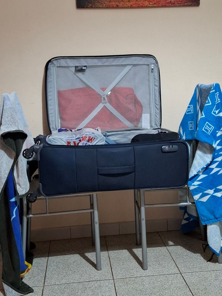 Blauer koffer auf 2 Stühlen liegend den aufgeklapptdn Deckel an die Wand dahinter gelehnt. Je ein Badetuch über eine der zwei Stuhllehnen im Koffer liegt Kleidung und in eine Plastiktüte verstaute Schuhe.