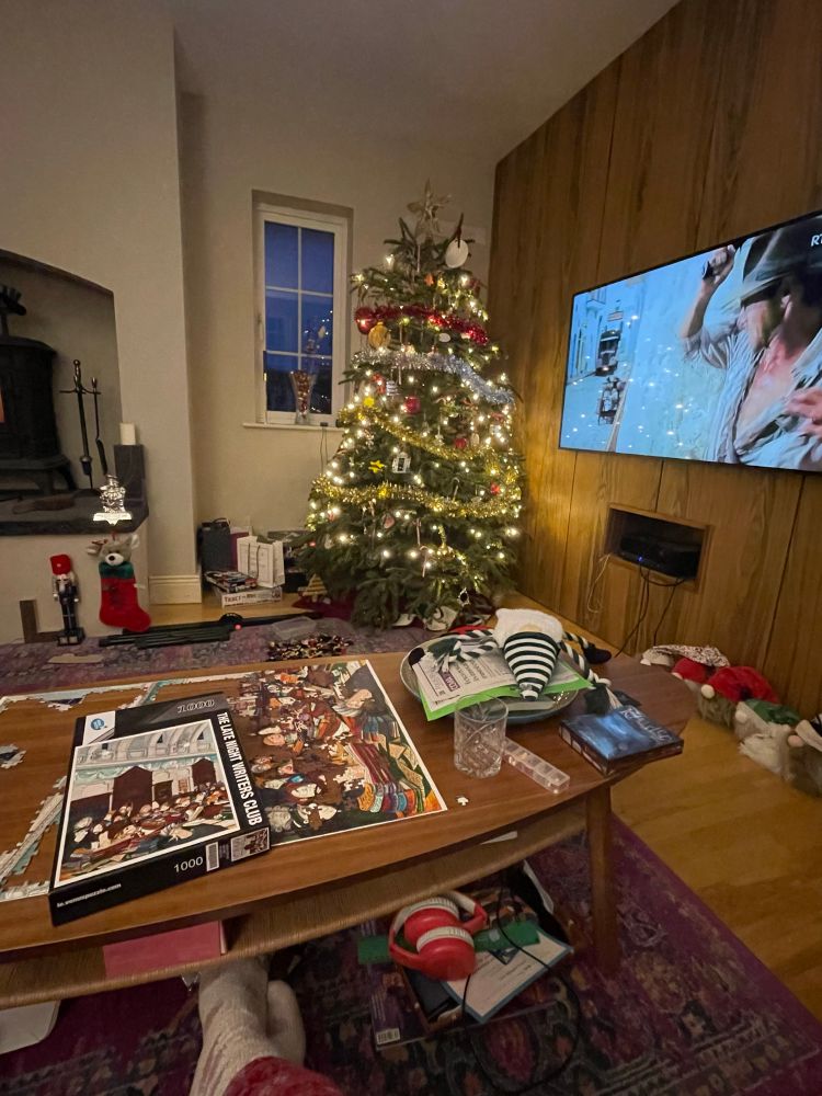 Foreground coffee table with jigsaw and Christmas detritus, tv screen on wall showing Indiana Jones movie and decorated Christmas tree in background