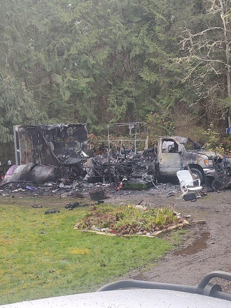 A class C RV gutted by fire. The heat of the fire has killed most of the plant life in a nearby garden. 