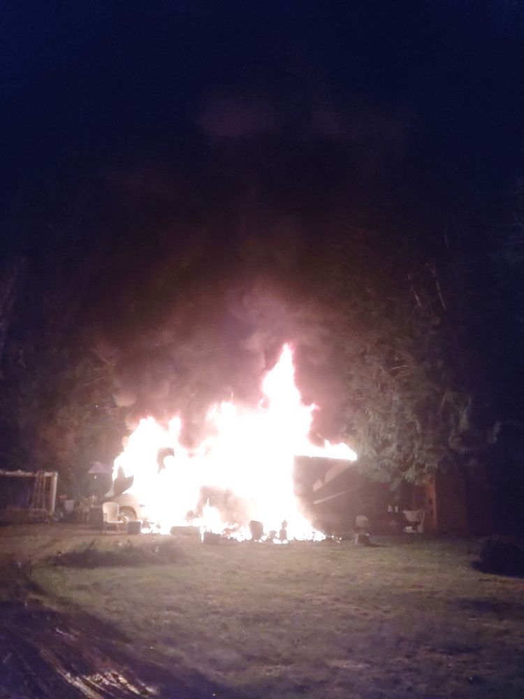 A column of fire envelops an RV in a pine forest late at night. 