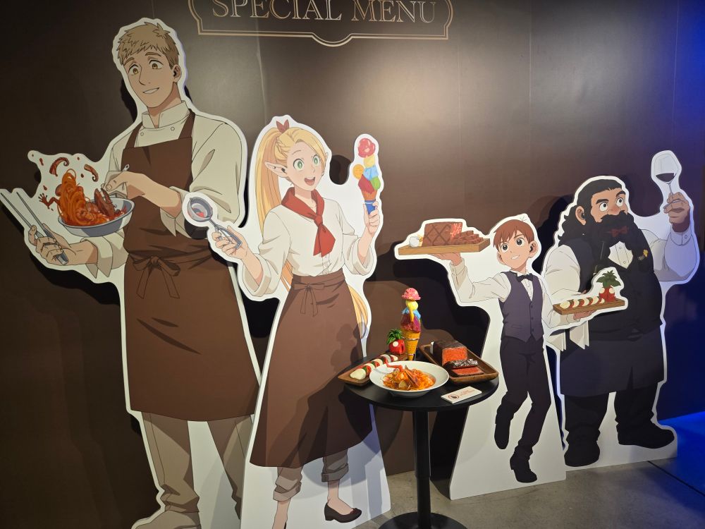 Cast of Delicious in Dungeon dressed as wait staff standees, with food sculptures in the middle.