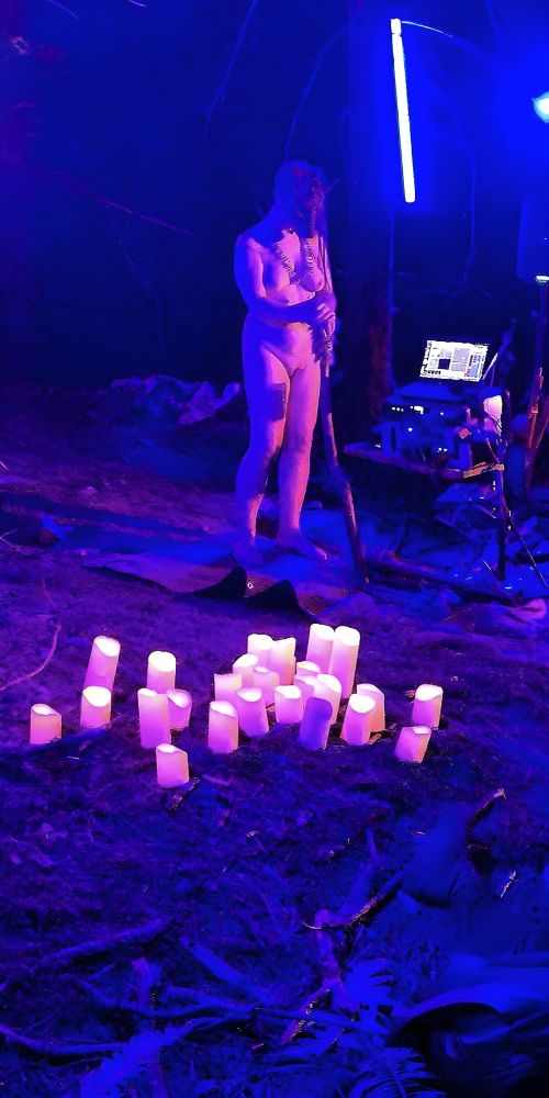 Image 2:
A slose-up of the Shimi, naked and masked performing under intense blue lighting, standing on stage surrounded by battery-powered candles. The figure wears a canine  mask and holds a staff, with visible body markings. They stand barefoot in the forest, illuminated by electronic stage lights, with audio equipment set up nearby. The scene evokes ritualistic and primal energy, blending performance art and forest ceremony.
Photo @seerendipity.bsky.social 

