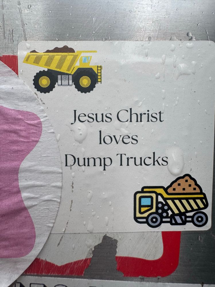 A small rectangular sticker on a metallic surface reads “Jesus Christ loves Dump Trucks,” printed in black serif text. Above and below the phrase are cartoon illustrations of yellow dump trucks carrying piles of brown dirt. The sticker is wet with raindrops and slightly weathered, giving it a street-art feel.