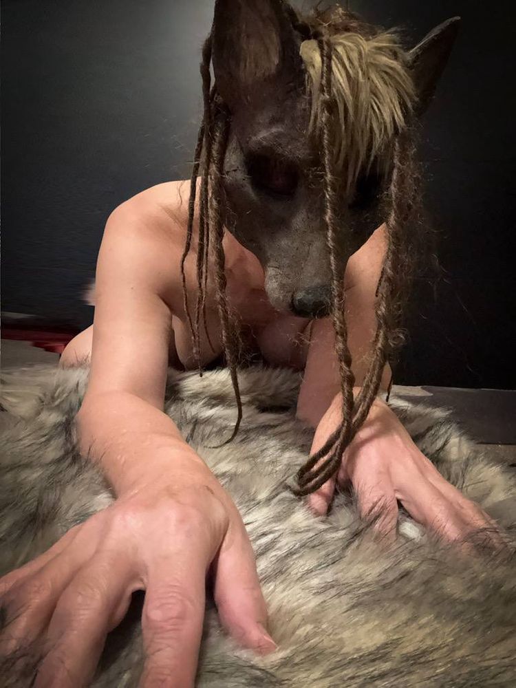 Shimi is wearing a realistic, canine-inspired mask is posed on all fours over a large, soft fur-like surface. The mask has pointed ears, textured detailing, and long braided strands of hair that hang down over the wearer’s face. Their hands are outstretched in front of them, fingers splayed into the fur, giving the scene an embodied, creaturely, immersive feel. The lighting is low and warm, with a dark background that emphasizes the mask and the tactile textures of skin, braid, and fur.