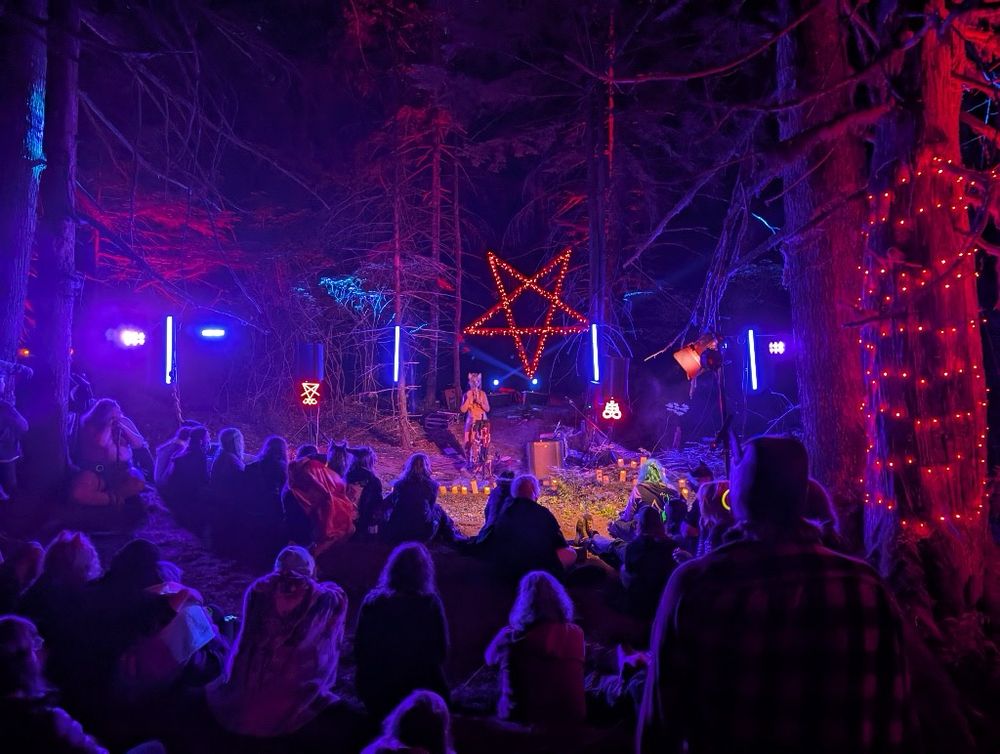 Image 1:
A nighttime forest performance scene lit in vivid purples, blues, and reds. A large crowd sits on the forest floor, facing a Shimi, performing naked and masked, standing on a small stage adorned with flickering LED candles. Behind the performer, a glowing red pentagram and various neon occult symbols are suspended between trees. Tall LED light bars emit blue light, casting an eerie yet magical ambiance. Trees are lit dramatically with red and blue spotlights, creating a mystical woodland theater atmosphere.
Photo: @wifewolf.bsky.social 