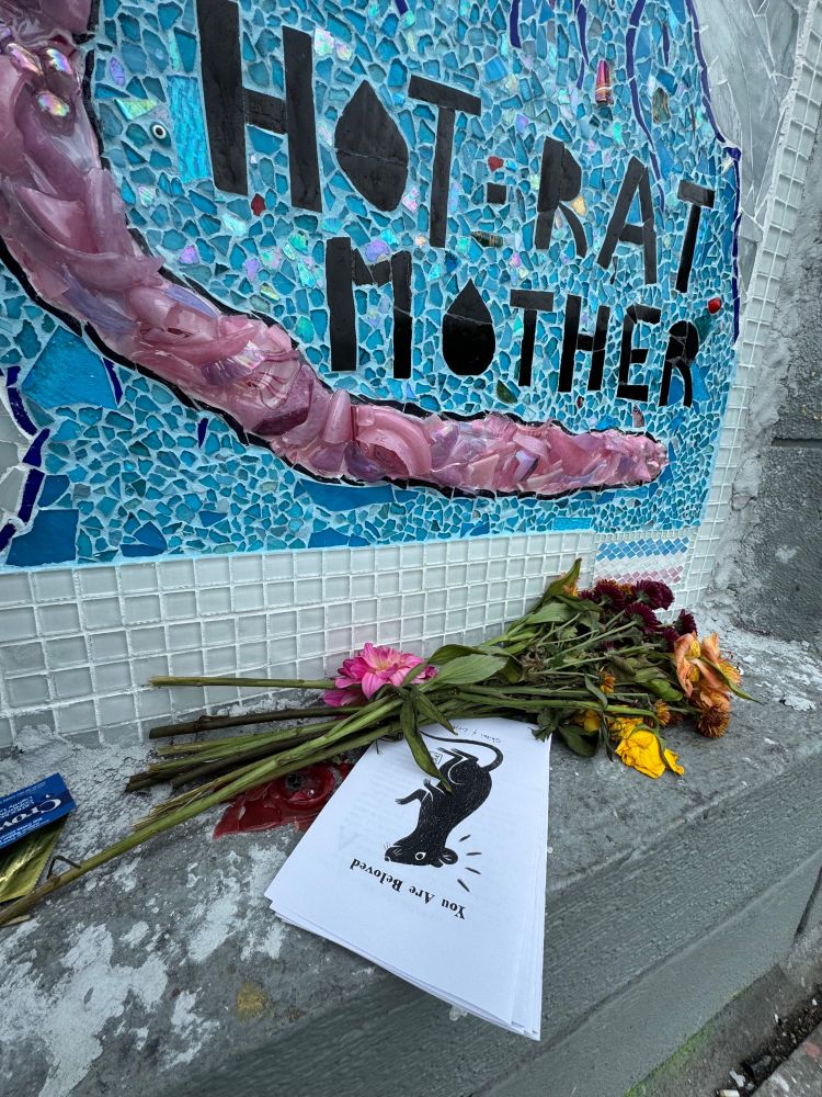 A small shrine built into a mosaic wall reading HOT RAT MOTHER in bold letters over shimmering blue tiles and a pink ceramic rat tail. On the ledge below, wilted flowers and a stack of zines titled You Are Beloved rest together, the cover showing a black rat with light lines radiating from its head like a halo.