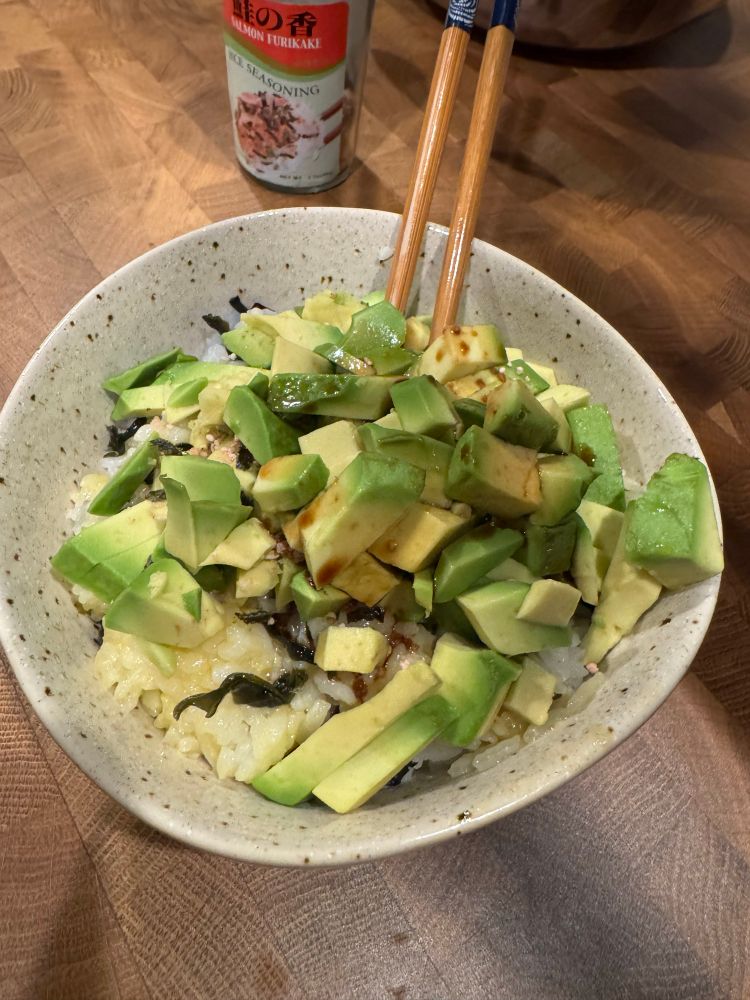 A ceramic bowl of rice topped with chunks of fresh avocado, seaweed, and a drizzle of sauce. A pair of wooden chopsticks rests on the rim of the bowl. In the background, there’s a container of salmon furikake seasoning on a wooden countertop.