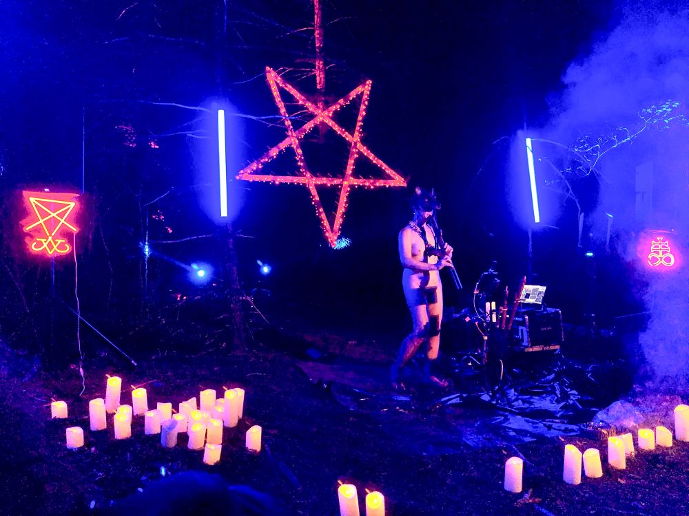 Image 3:
Another wide view of Shimi performing on stage during the night-time forest ritual. This wolf-masked figure plays a flute amid glowing candles, while a large red pentagram and other neon symbols illuminate the space. Blue vertical LED lights frame the scene, and fog or smoke adds depth and mystery. The ground is covered in forest debris, contributing to the raw and elemental mood.
Photo @shade-the-ashclad.bsky.social 