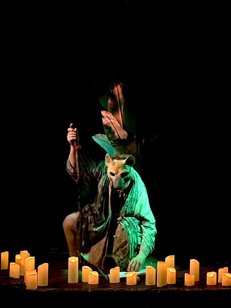 The same performer (Shimi) kneels in a different pose, centered within a semicircle of glowing candles. They wear the same wolf mask and long, flowing robe, now illuminated by a cooler green light. Their left hand rests on an open book on the floor, while their right hand holds the tall staff upright. The backdrop remains dark, with the same dramatic leaf shapes rising behind them. The scene feels reverent, theatrical, and ritualistic—invoking the presence of a sacred beast or shamanic figure.