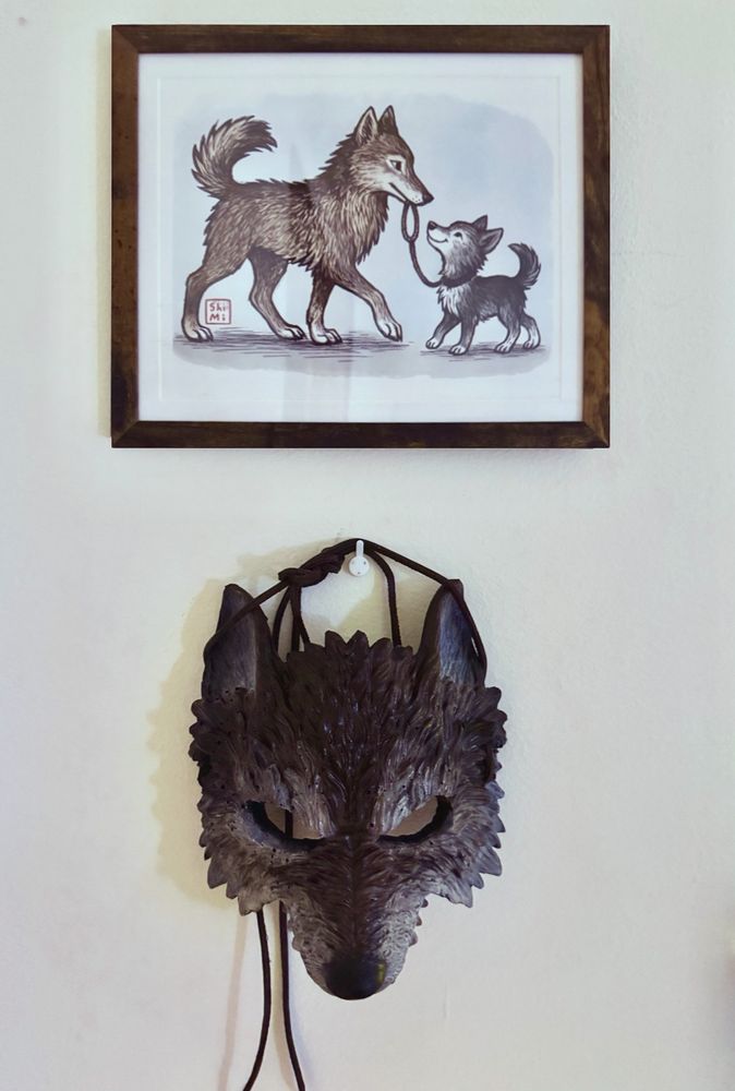 A photograph of a wall with two art objects. The upper picture shows a stylized illustration of two anthropomorphic figures. On the right is Shimi, a slightly smaller character with gentle features. On the left is a larger anthropomorphic coyote boy, who Shimi is dating. The two appear close and affectionate. 

Below this frame, mounted on the wall, is a sculpted wolf mask with pointed ears and a long snout, pale in color and expressionless, watching outward from the center of the image.