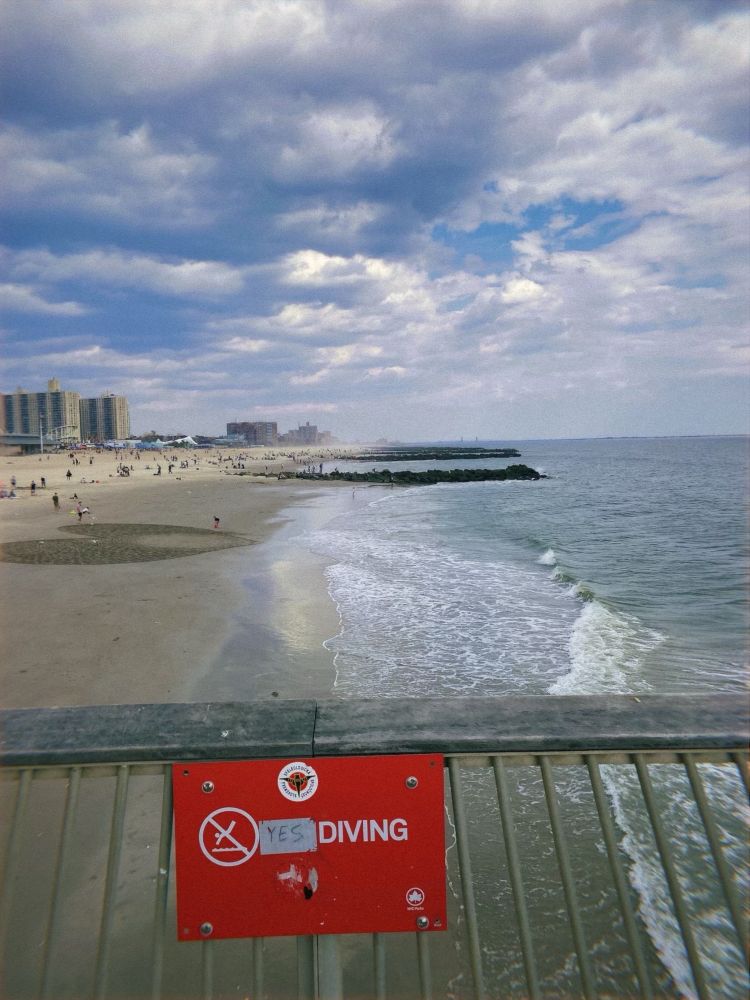 A side view of Coney Island beach. A red warning sign has a makeshift alteration that makes it say “YES DIVING” instead of “NO DIVING.” In the distance a large heart is carved in the sand. 