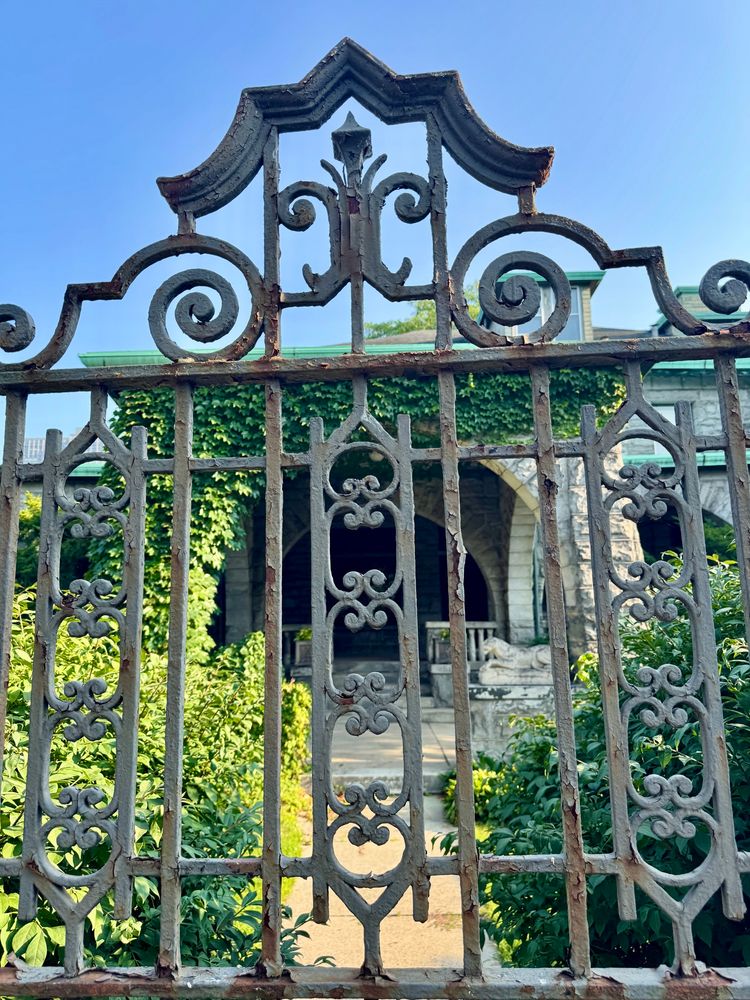 Closeup of a decorative, rusted iron gate in front of 803 W. Hutchinson St. in Chicago’s Uptown neighborhood. Green ivy vines cover the archway entrance of the home behind the gate. A statue of a resting lion is on a pillar near the door.