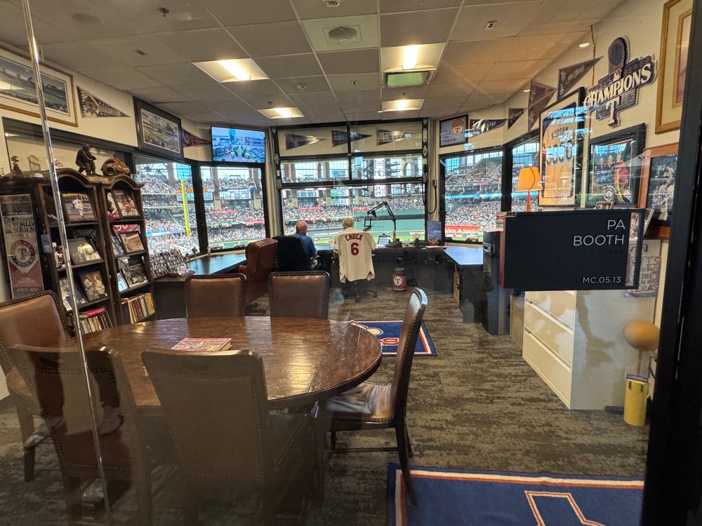 View inside a room labeled PA booth from behind a soundproof glass window. Fans can watch Rangers public address announcer Chuck Morgan do his job from the booth. A small brown conference table is placed near the glass window. The walls are lined with pennants, framed photos and cases of baseballs and other memorabilia. At the doorway is a large blue rug with a blue 1980s-era Rangers logo.