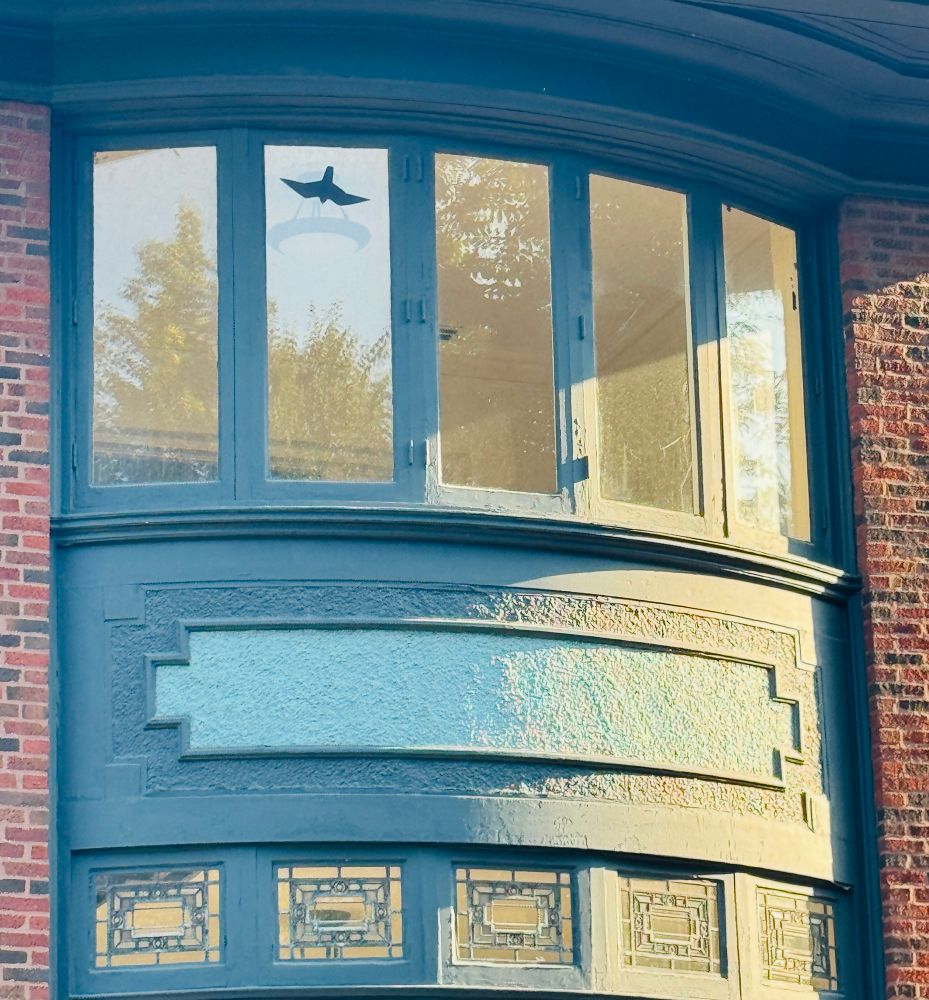 Closeup of a Prairie-style green metal window with decorative panes at the bottom and brick surrounding it on either side. On one pane near the top is a black sticker of a small bird. The room inside is painted beige and is completely empty. The house at 817 W. Hutchinson St. in Chicago was designed by George Maher.
