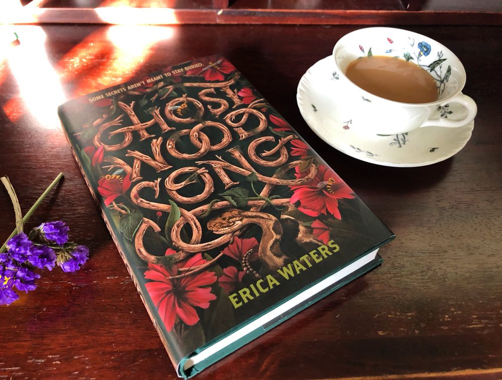 Ghost Wood Song’s hardcover lies on a table between a cup of tea and dried purple flowers. The cover features a large stylized title that looks like it is made of wood, as well as azaleas, wasps, and a rattlesnake. 