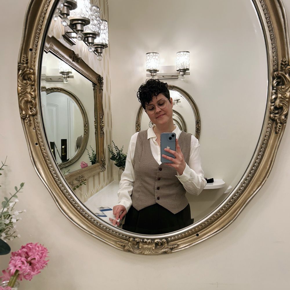 The photo shows a white nonbinary person wearing a vest and taking a picture in a mirror. 