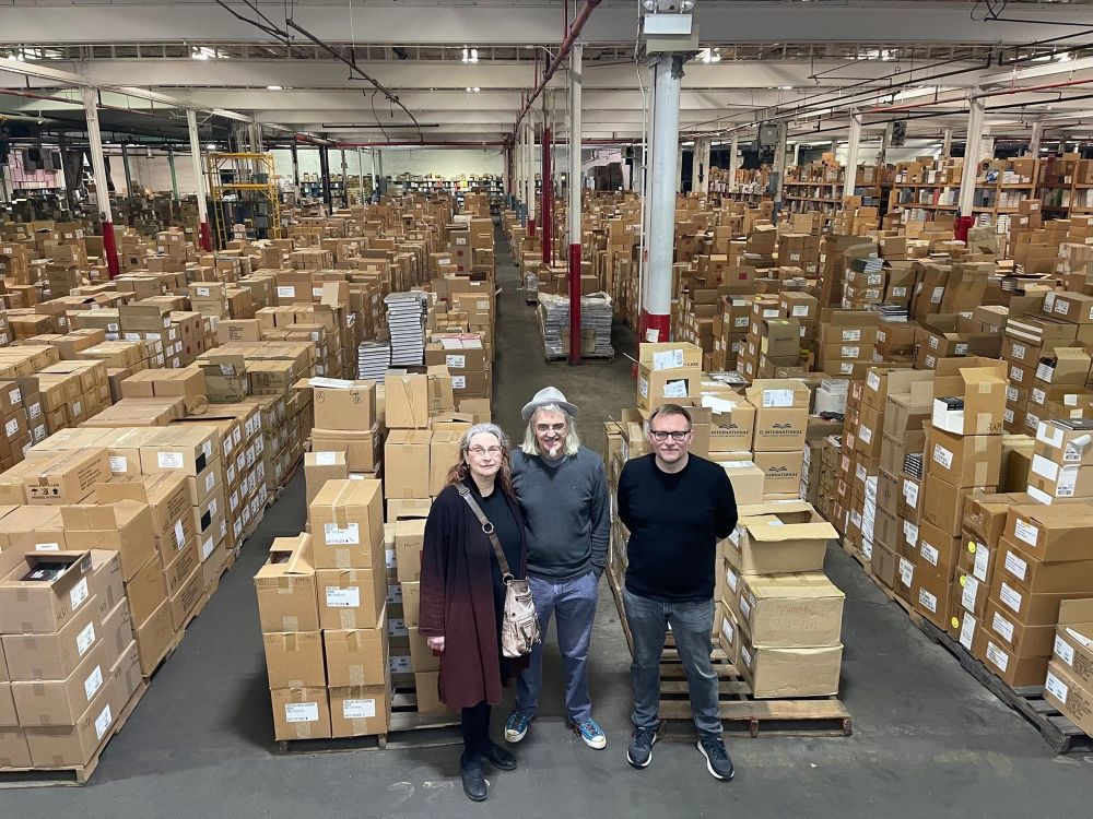 Audrey, Eddie, and Ken standing in an enormous warehouse in front of thousands of boxes of books. We are all smiling!