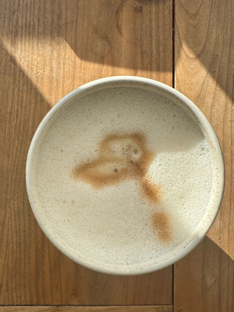 Ein Becher Milchkaffee steht auf einer hölzernen, halb von der Sonne beschienenen Tischplatte. Fotografiert aus der Vogelperspektive. 