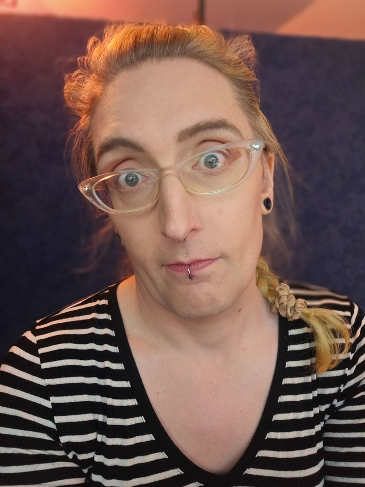selfie of a white trans woman with blonde hair and transparent cateye glasses staring into the camera. she is wearing a black and white striped shirt and has her hair in a braid that is visible over her left shoulder. she has been clenching her jaw for so long that she isn't entirely certain how one would open one's mouth. 
