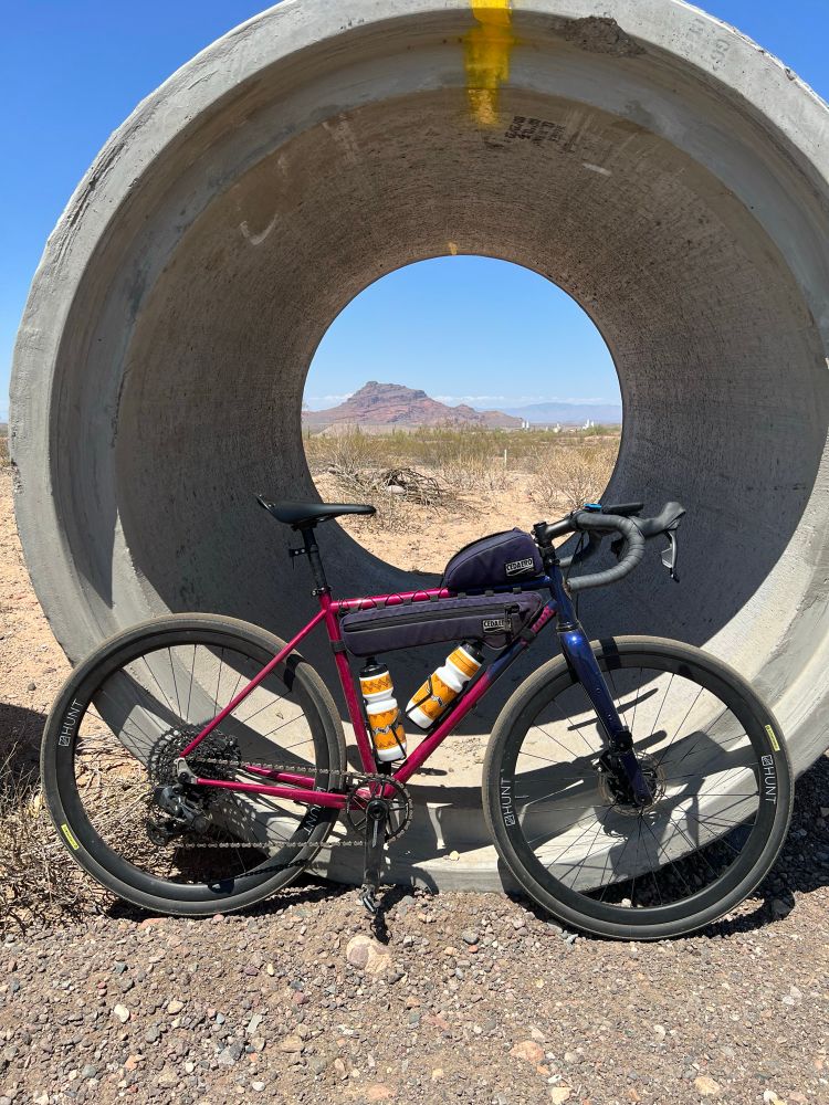 a gravel bike leaning against a large section of concrete pipe. in the background is a mountain that can be seen through the pipe. 