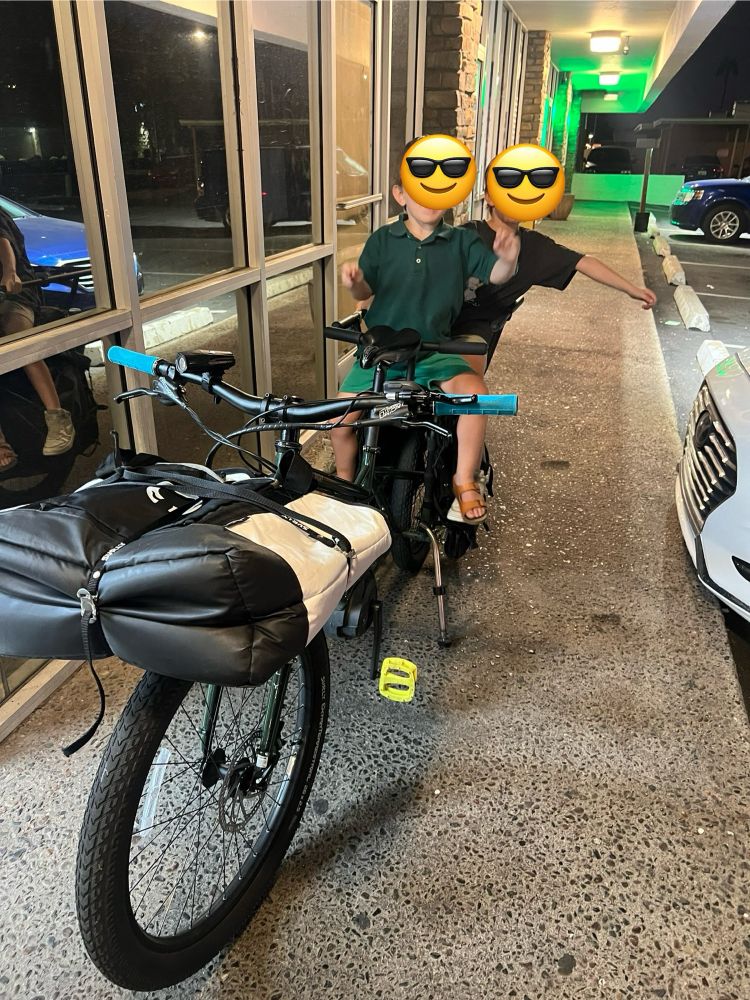 two kids on the back of a cargo bike. strapped on the front rack of the bike is a reusable grocery bag with two pizzas inside
