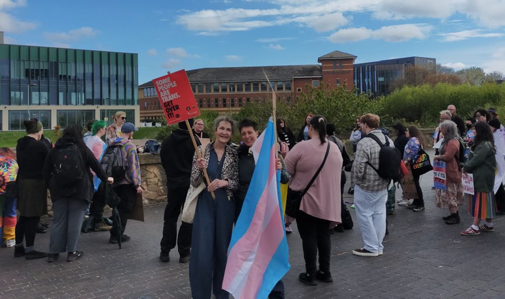 A trans woman and a non-binary person protest in Middlesbrough. They carry a placard and a trans flag, and are surrounded by other protestors.