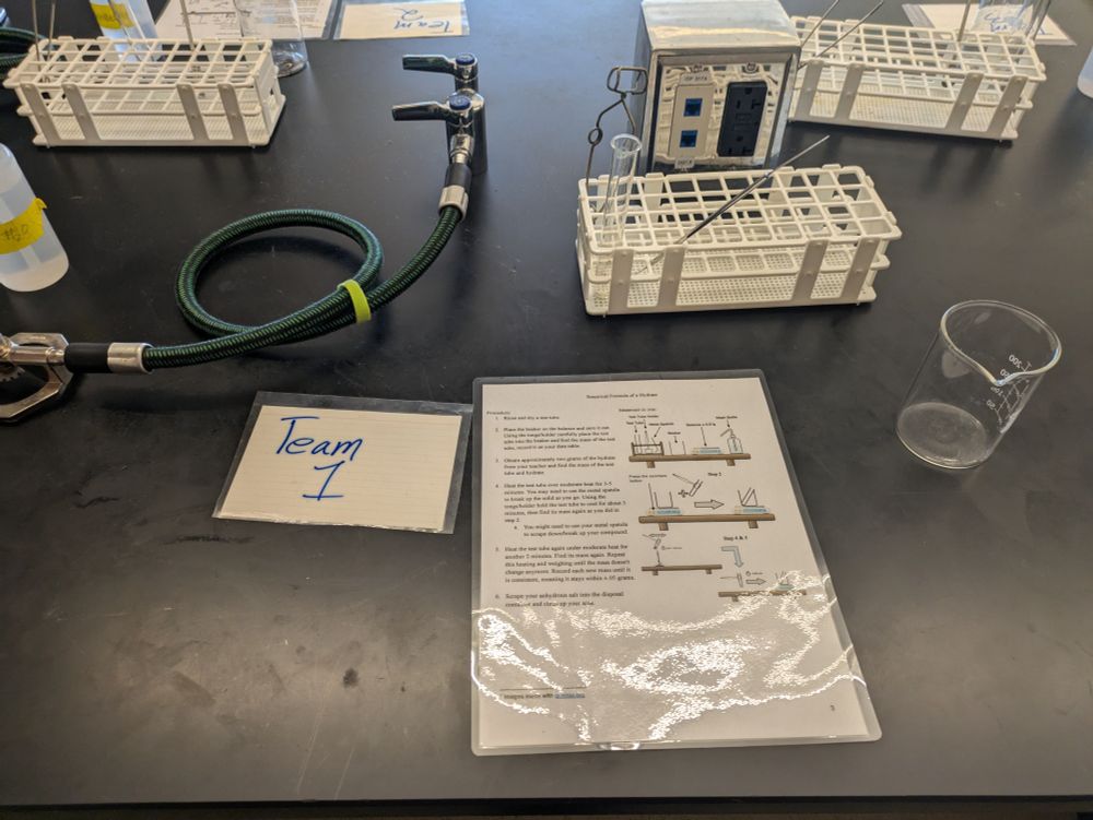 Lab table setup with labeled "Team 1" sign. Includes a green hose, beaker, test tube racks, and a procedure sheet. Clean, organized workspace.