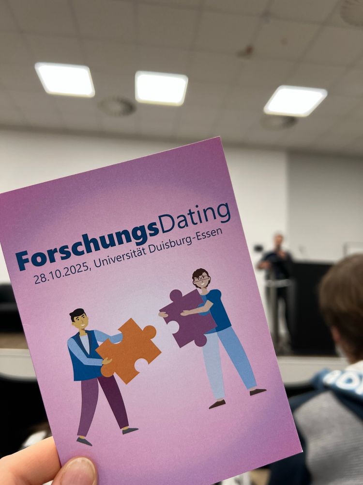 Card with text “ForschungsDating, 28.10.2025, Universitär Duisburg-Essen” and Two people with puzzle pieces; in the background stage with person talking