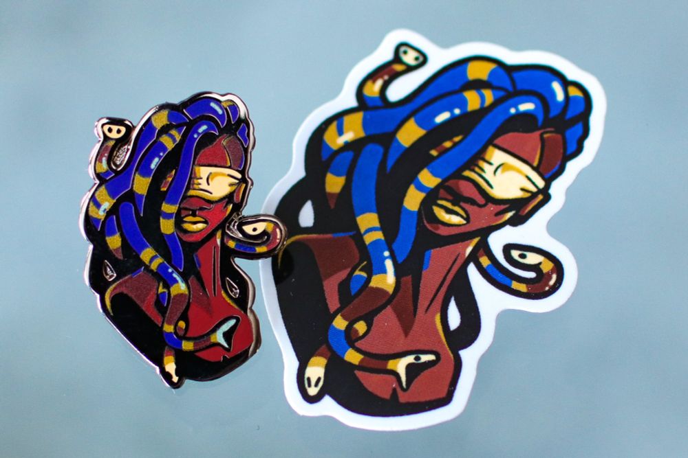 Picture of the medusa pin and matching sticker from Kill Every Monster. They are busts of a brown-skinned woman, blindfolded, with a head of coral snakes that resemble dreadlocks with gold bands.