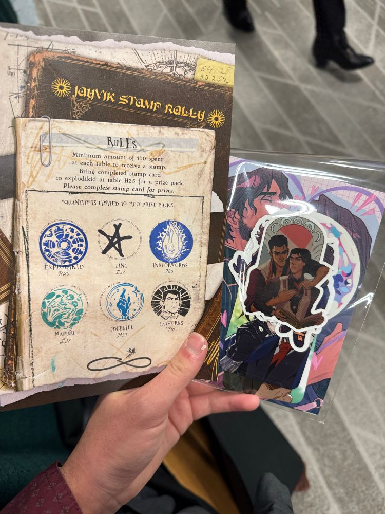 Hand holding up a filled out stamp punch sheet titled “Jayvik Stamp Rally” and a bag of Jayvik prints and stickers.