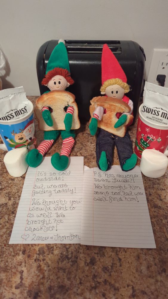 Two elves sit against a toaster with their arms through a slice of toast. On each side is a Christmas mug, with a hot chocolate package and a giant marshmallow. In front of them are 2 pages of curly script reading "It's cold outside but we are getting toasty! We thought you would want to as well! We brought hot chocolate!

Love Lance and Thornton. 

P.S. has anyone seen Lucas?! We brought him some too!"