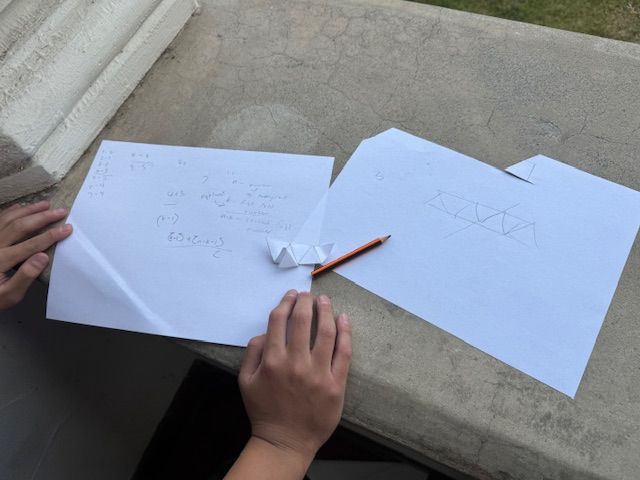 A pencil, paper, and a partially folded piece of paper folded into a strip of equilateral triangles.
