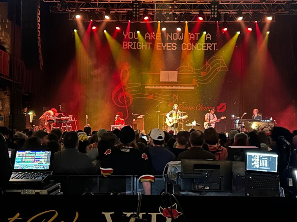 Bright Eyes on stage with a banner saying “you are now at a bright eyes concert” 