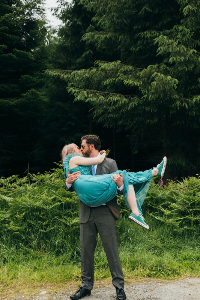 Man holding a woman in a green wedding dress beside a forest while they kiss. 