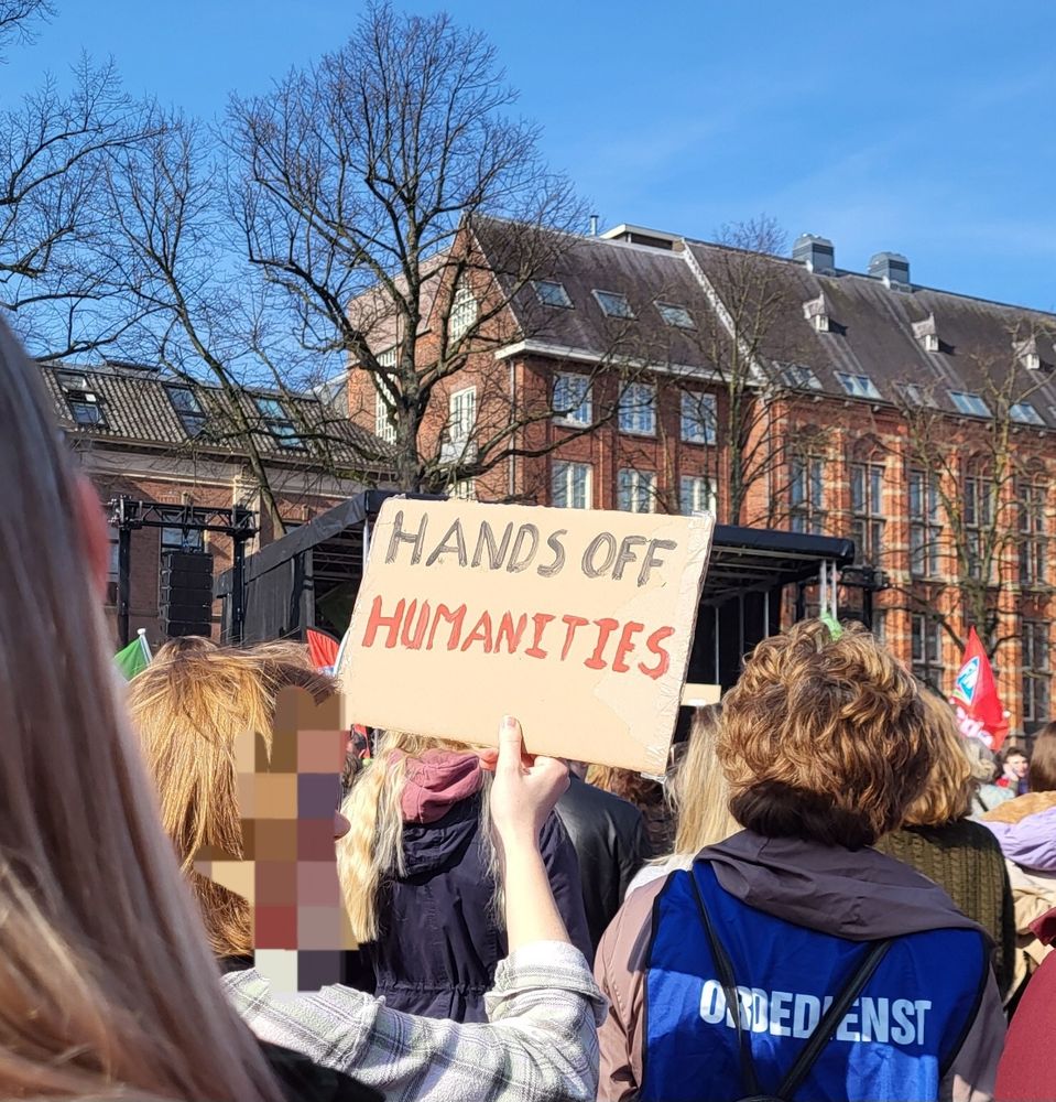 People standing in a demonstration, seen from behind. A person has a sign that says: Hands Off Humanities 