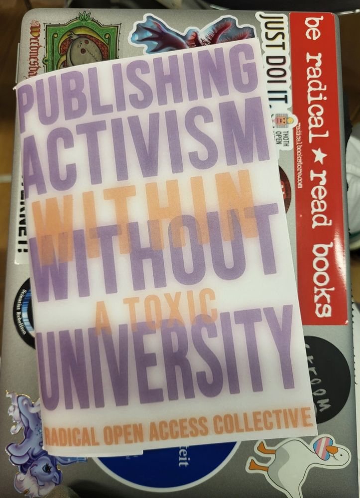 A booklet: Publishing Activism with/without a Toxic University on top of a laptop with many stickers, among them one that says: be radical, read books. And a white goose holding a heart with the trans flag colours.