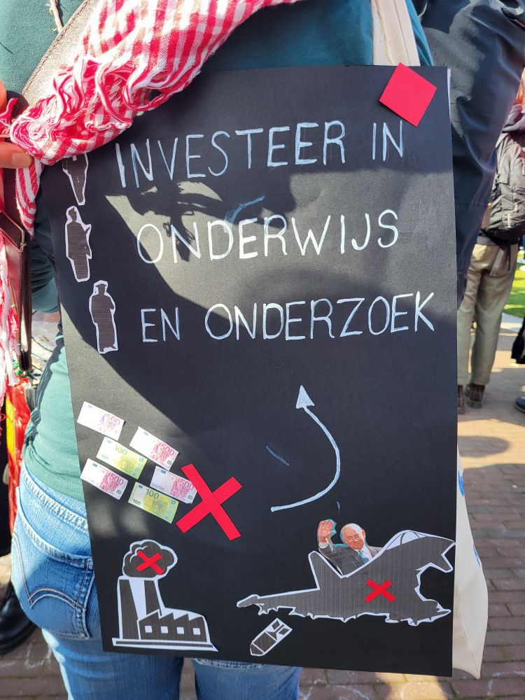 A poster collage in black paper with white letters and cutouts from magazines. The text says Investeer in Onderwijs en Onderzoek. There are euro bills going into polluting factories  and fighter plane commanded by Benjamin Netanyahu, these are crossed with red and an arrow indicates the money should go to Education and Research