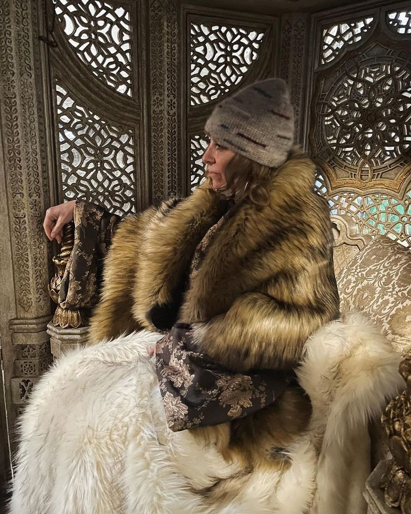 Picture of Sharon Gilham sitting in Siuan's exquisitely carved palanquin, wearing furs and a luxurious coat. She has a grey beanie on.