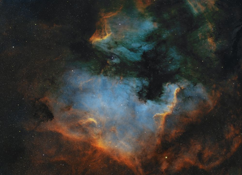 The North American nebula reclines below the Pelican nebula. Dark red-orange swirls of dust are present, with hues of blue and green above. A blanket of stars is scattered throughout. 